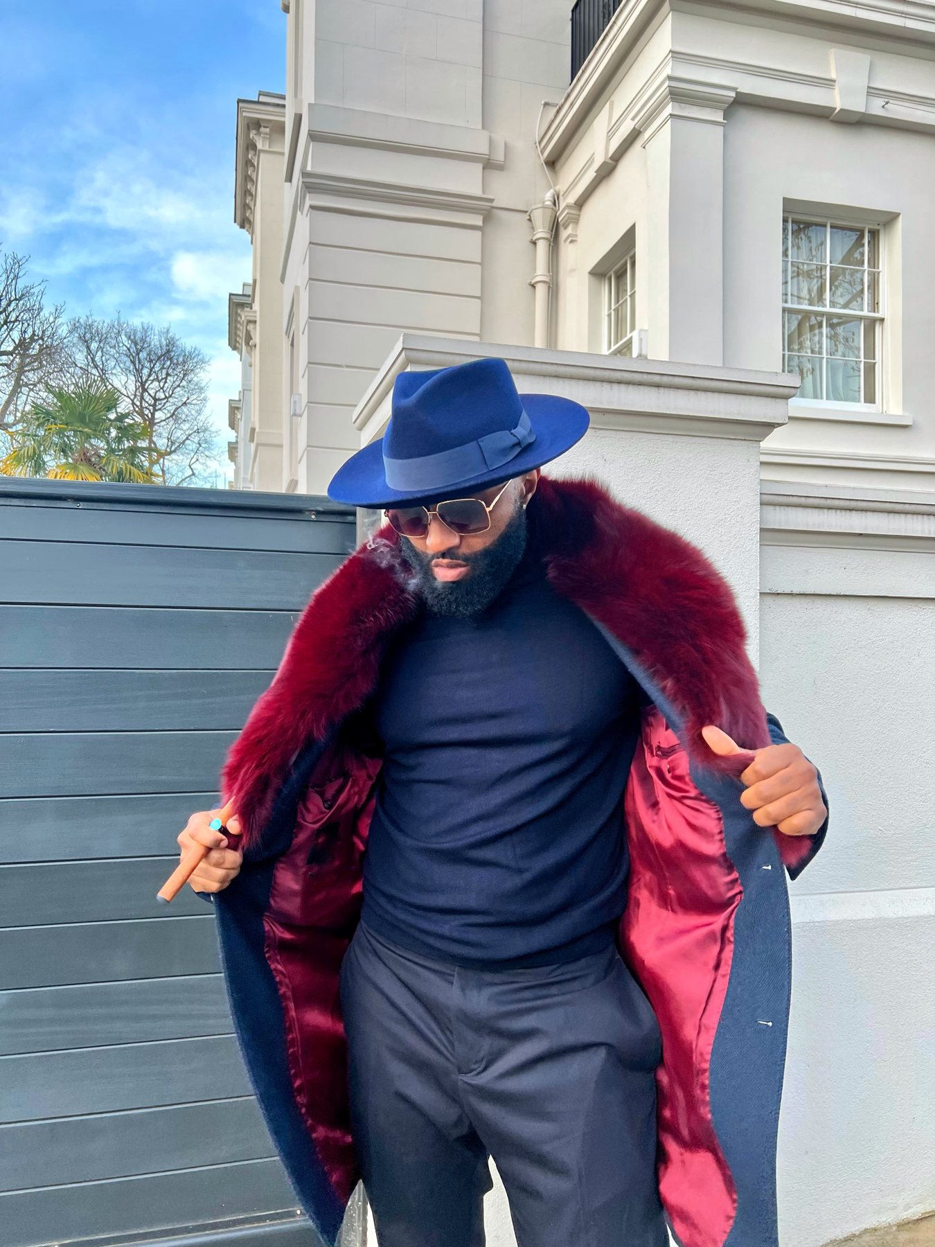 Man in navy overcoat with burgundy fox-fur collar and navy fedora outside a white London townhouse
